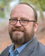 Dr. Feodor Gostjev smiling with short balding reddish brown hair, mustache and beard, wearing black rim glasses and a gray blazer over a light blue button up shirt